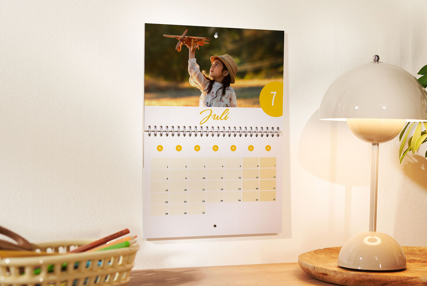 Op een wandkalender staat een vrolijk kind afgebeeld dat een speelgoedvliegtuig vasthoudt. De maand juli is gemarkeerd.