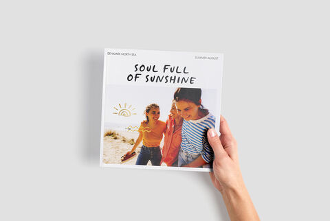 Une personne tient un album photo CEWE avec le texte 'Soul Full of Sunshine' devant un fond estival.
