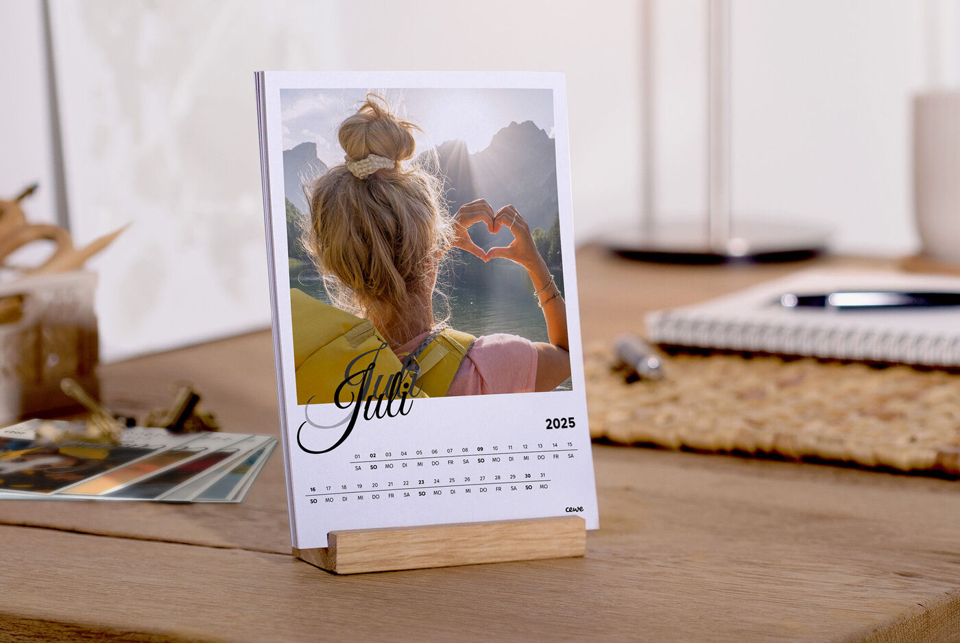 Une personne aux longs cheveux forme un cœur avec ses mains devant un paysage pittoresque, un calendrier personnalisé sur la table.