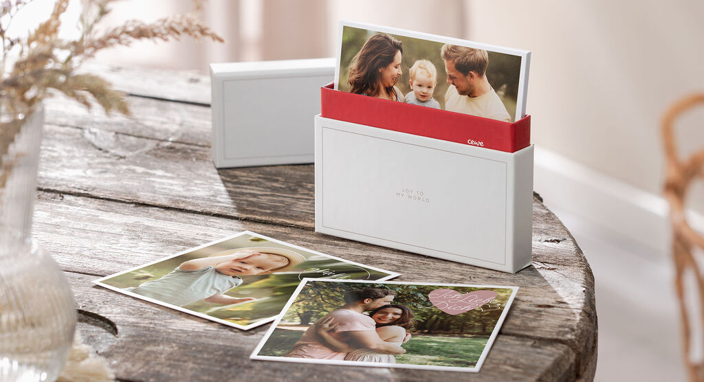 Cartes de vœux CEWE dans une boîte blanche sur une table en bois, entourées d'autres cartes photo.
