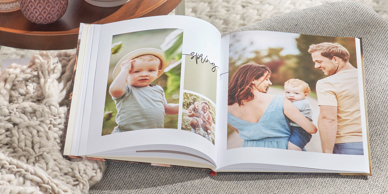 Op een grijze bank ligt een CEWE FOTOBOEK. Op de cover zie je een foto van een gezin en de tekst "Moments 2021". Aan de linkerkant staat een tafel met theelichtjes en een plant.