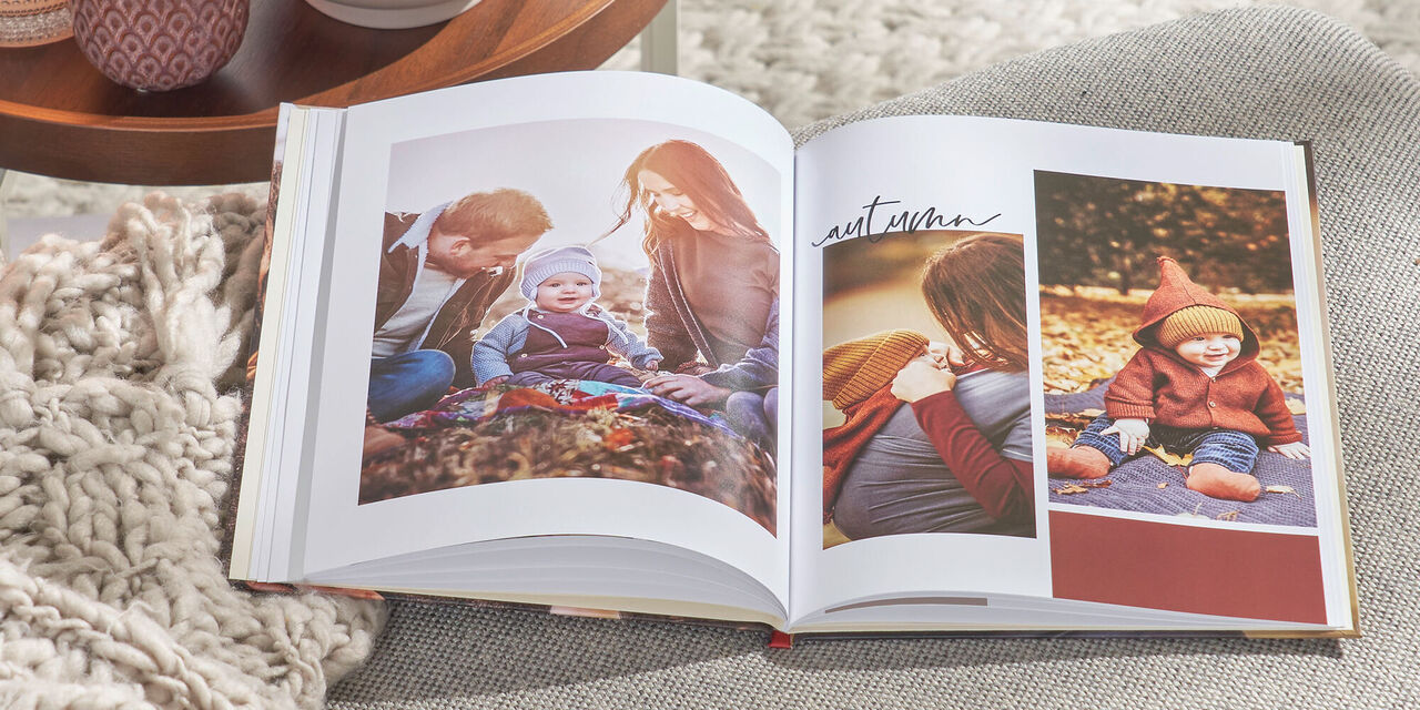 Op een grijze bank ligt een CEWE FOTOBOEK. Op de cover zie je een foto van een gezin en de tekst "Moments 2021". Aan de linkerkant staat een tafel met theelichtjes en een plant.